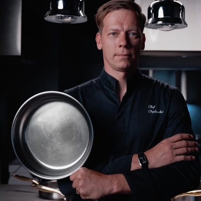 A man in a black chef's jacket stands in a dark kitchen setting, holding a large stainless steel sauté pan. The chef's jacket has white embroidered text on the left chest that appears to read 'Chef N.