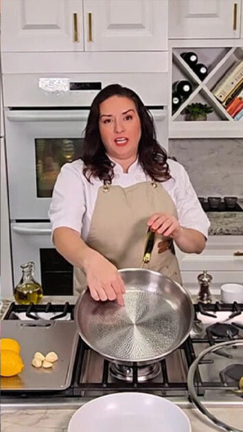 A woman in a white shirt and beige apron stands in a kitchen holding a stainless steel frying pan with a gold handle over a gas stove.