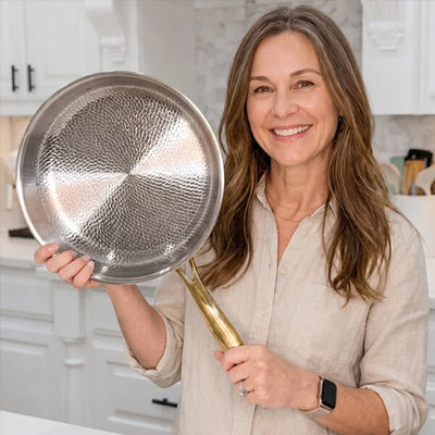 A smiling woman in a light beige linen shirt holds up a hammered stainless steel frying pan with a brass handle in a bright, modern kitchen setting.