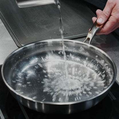A Titanium frying pan with a textured, dimpled interior surface is shown on a stovetop with water being poured into it. A hand is holding the handle, and a dark stovetop surface is visible in the background.